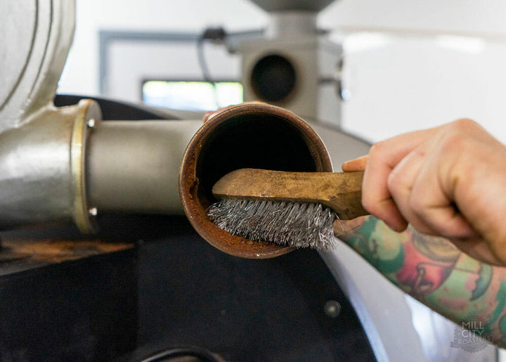 There is a hand coming out of the right lower corner holding a brush with metal bristles and a wooden handle. The brush is scrubbing coffee residue off the inside of a vent coming off of the coffee roaster.