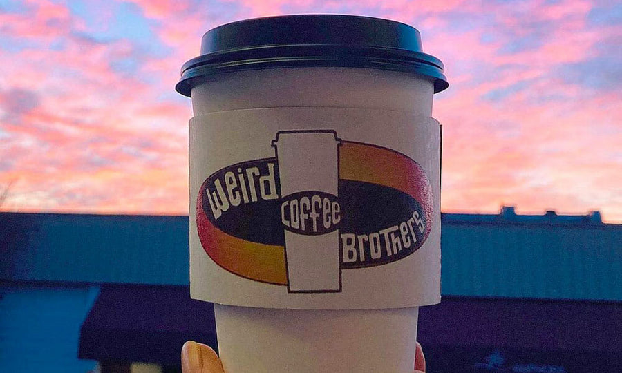 A Weird Brothers Coffee to go cup is held up in front of a sunset