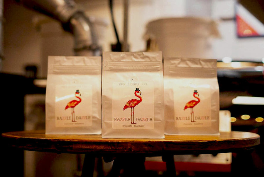 Three bags of coffee positioned on a small round table in front of a roaster. On the bags there is a pink flamingo with text at the bottom of the label.