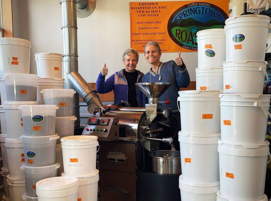 A couple stand behind a roaster smiling at the camera. They are surrounded by buckets of roasted coffee.