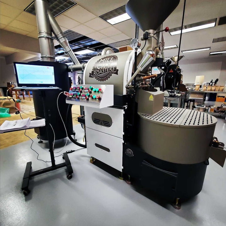 A couple standing together in front of a small roaster. Below them there are bags of green coffee beans.