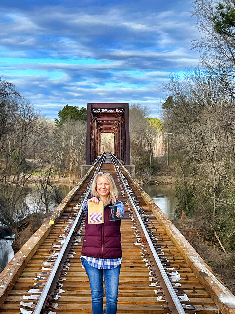 Someone standing in the middle of train track holding out a bag of coffee beans and a cup of coffee. They appear to be surrounded by nature.