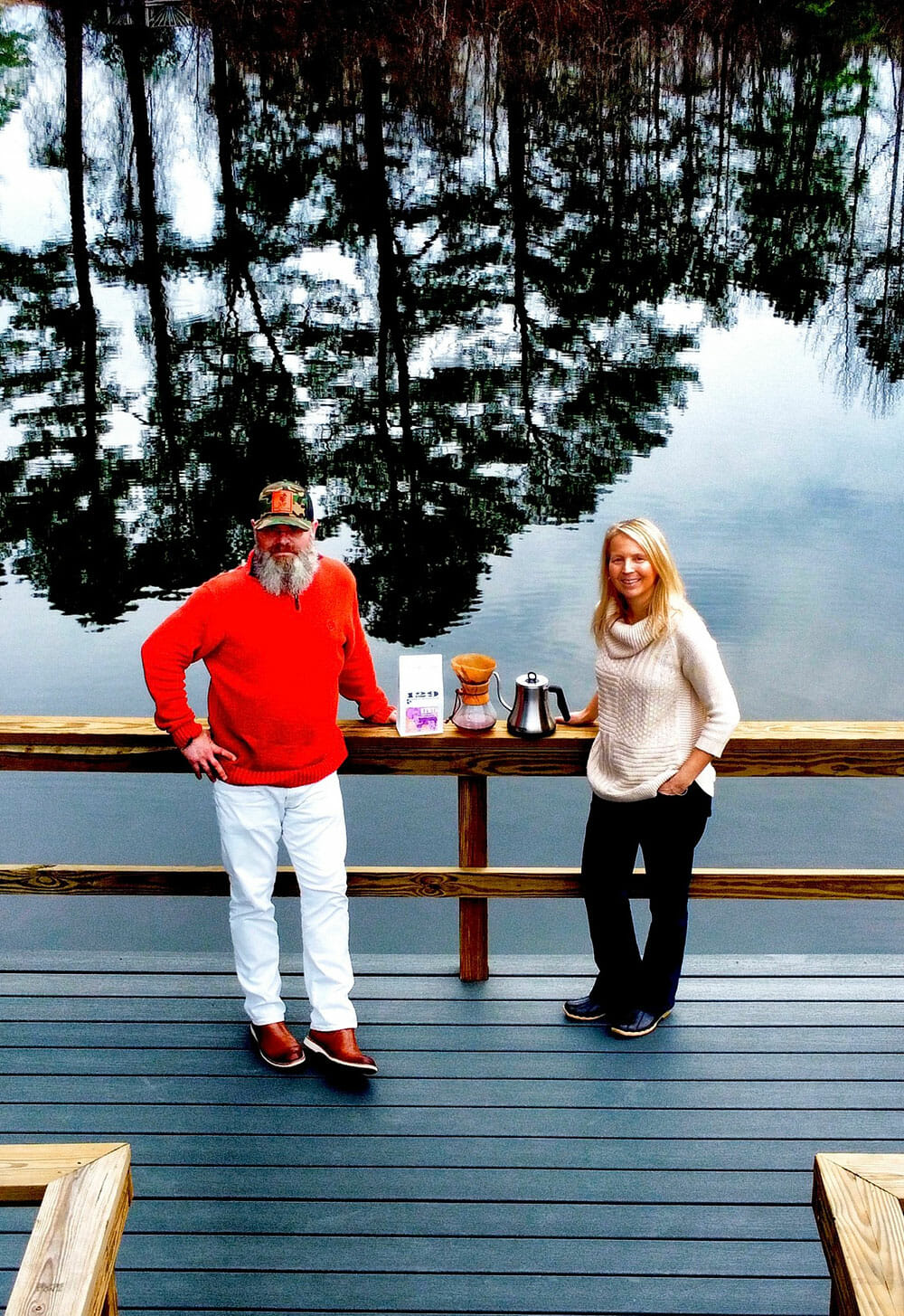 A couple stands against wooden railing with coffee and brewing equipment between them. Behind them the lake reflects the shadow of trees.