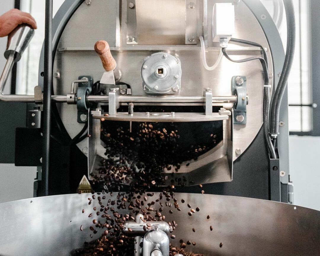 The front of a roaster with the door open. Coffee beans are spilling out of the roaster into the cooling tray.