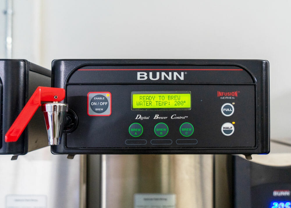 The top of a commercial batch brewer. It is black with a small rectangle screen in the middle. Below the screen are three brew button options that read Brew A, B and C. To the far right there two brew options for either a full batch or half batch. To the far left, there is another round button for turning the commercial brewer on and off.
