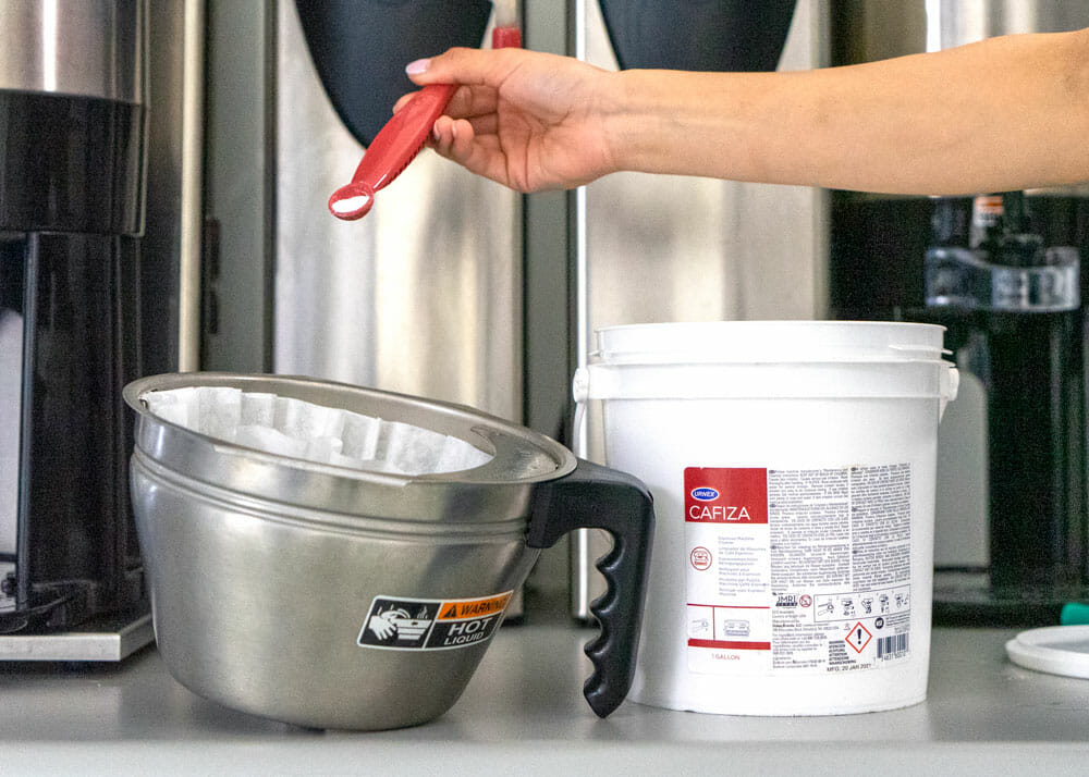 A coffee brewer basket is sitting on a grey counter with a white paper filter inside of it. The handle to the brewer is black and is facing the right side. Next to the handle is a bucket of cleaning powder with a red label on the front. There is a hand about the brew basket and cleaning container holding a red scoop. The scoop is dispensing powder into the brew basket.
