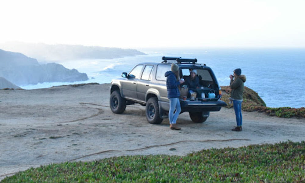 Three people drink freshly brewed coffee from the back of a truck on a cliff overlooking the ocean