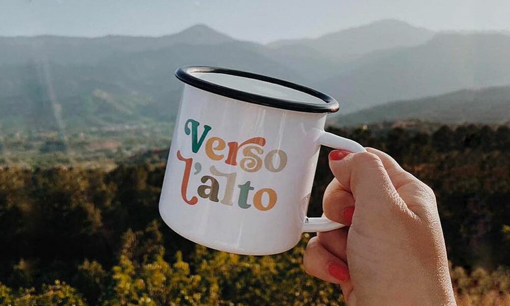 A hand is holding a Verso L'Alto branded cup with a bright and sunny mountain range in the background