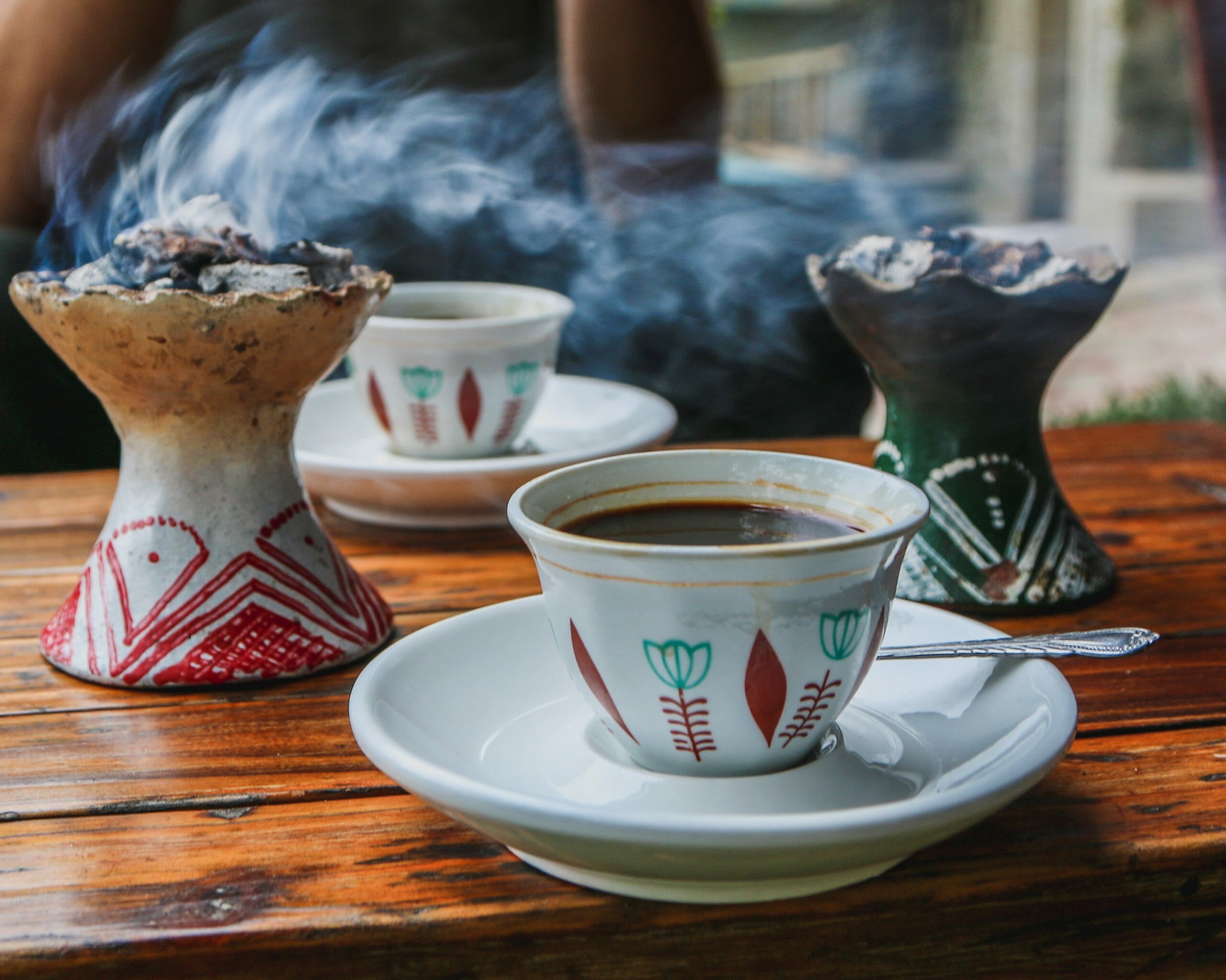 Two white porcelain mugs each sitting on a white saucer. There are green and red patterns on the outside of each mug. To the left, is a ceramic incense burner with red patterns on the side. To the right, there is another ceramic incense holder that is green with white patterns on the outside. Both incense holders have smoke coming from them.
