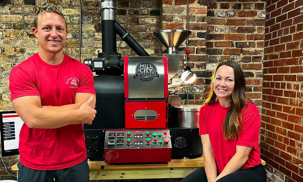 Two people standing on each side of a coffee roaster between them. They are both wearing red shirts to match the red colored roaster.
