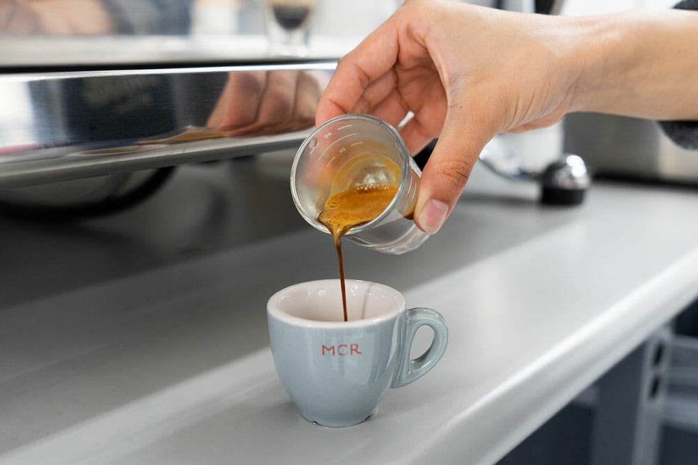 Someone is pouring coffee from a measuring shot glass into a small blue espresso cup. On the cup, the letters "MCR" are printed in red in front of the espresso cup.