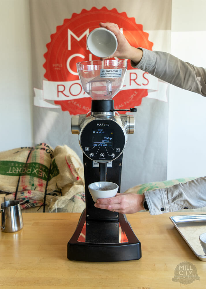 A coffee grinder is in the middle of the image. Above the beans hopper, a person is tilting an empty white cup. Below the doser, ground coffee is pouring into another cup that the same person is holding in their other hand.