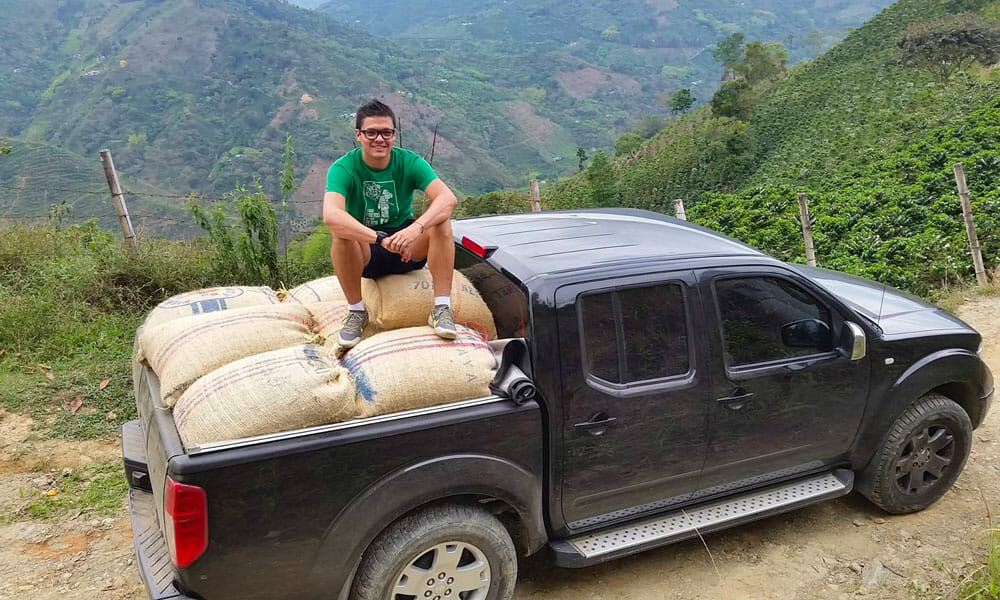 Someone sitting on burlap bags of green coffee in the back of a truck. The truck is parked on a dirt road with rolling green mountains in the background.