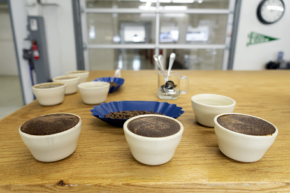 Cupping bowls filled to the top with coffee at the edge of a wooden table.
