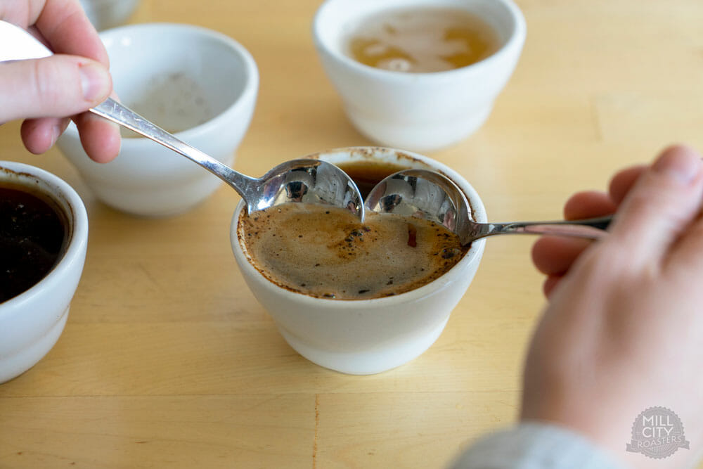 Someone holding two spoons together to scrape of the crema of the coffee in the cupping bowls.