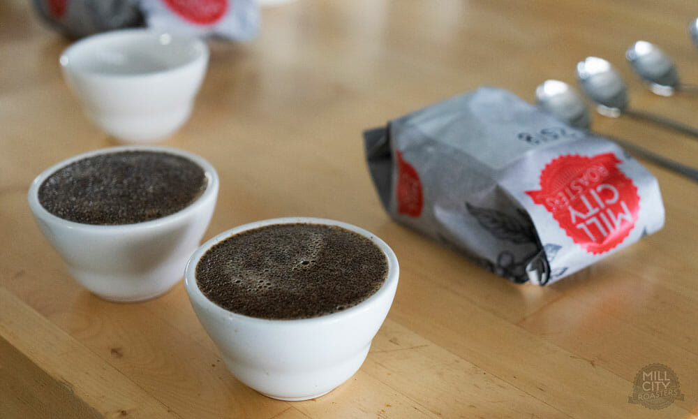 A few small white bowls sit on a wooden table with bags of coffee, and spoons laid out as well. In two of the bowls, to the left, there is brewed coffee in them with a formed crust of grounds on top acting as a barrier to the liquid below.