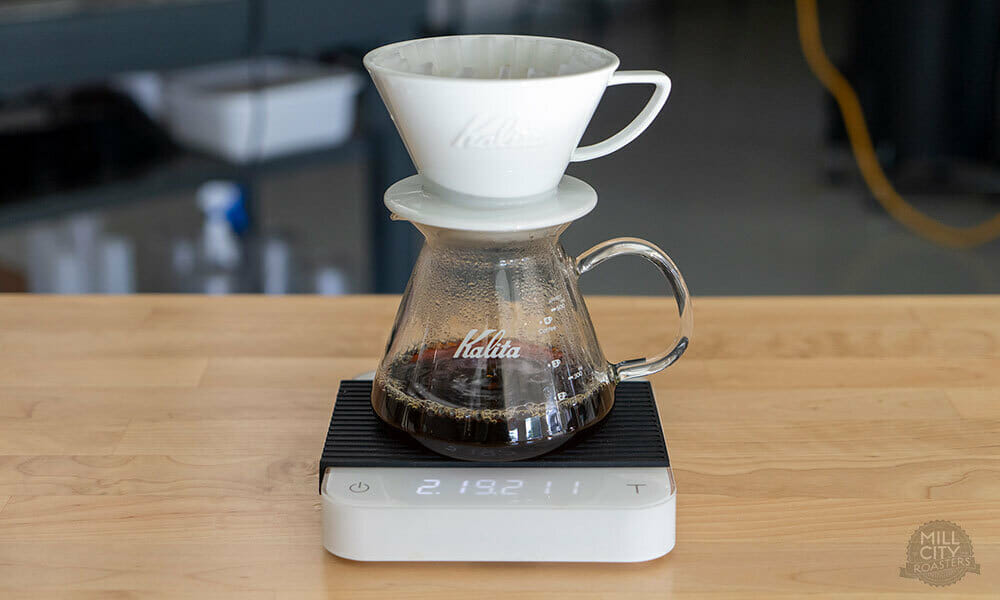 A pour-over brewing system on top on a white scale white a black non-slip sleeve. Coffee is draining while a timer counts down on the scale.