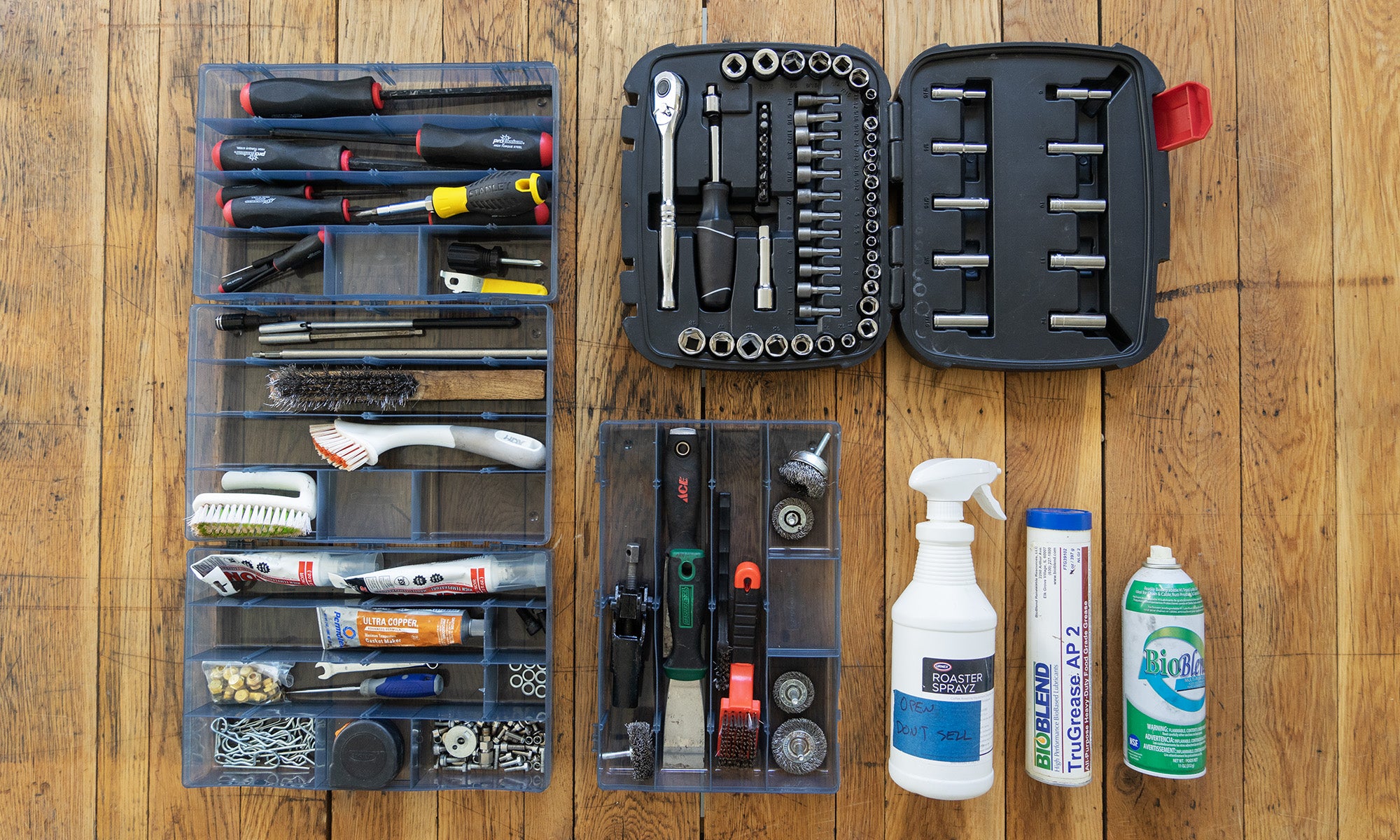 A wooden table with tools, sprays, brushes, and grease. In the top left corner, there is an assortment of screw drivers and allen keys. Below that there are soft bristle brushes and to the right of that there are paint scrapers. In the far right corner a tool box lies open with a set of nuts and bolts with a wrench. At the far bottom to the right, there is spray and grease used for roaster maintenance.
