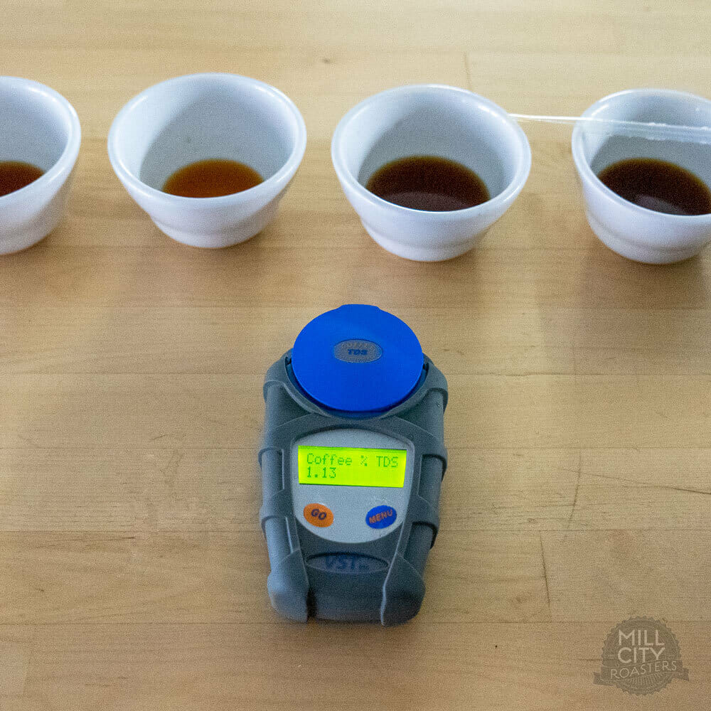 Small white bowls lined out on a wooden table with coffee in them. In front of the bowls there is a commonly used measuring tool for brewed coffee. It has a moveable blue flap on top with a yellow LCD display.