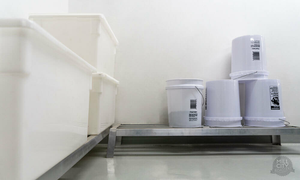 To the left of the image there are off white, transparent totes lined up against the wall. Straight ahead, there several white buckets stacked on top of each other. The the left of the stacked pile there is one bucket face up with coffee inside of it. All of the containers are sitting on small metal shelves that help elevate things.
