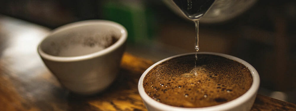 On a dark wooden table there are two small white ceramic bowls holding brewed coffee. At the surface of the bowls there appears to be coffee grounds creating a barrier above the liquid beneath. From the top of the image there appears to be a kettle pouring water into the bowl to the right.