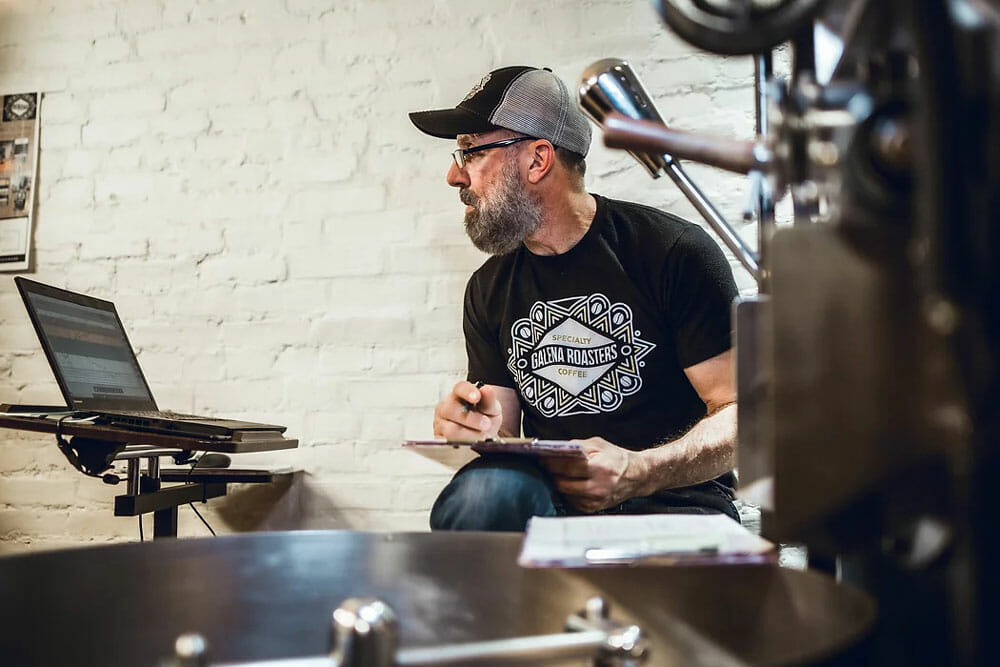 In the forefront on the image is a coffee roaster. Right behind it is a man wearing a black shirt and glasses holding a clip board in his lap as his sits next to the roaster. He's looking to the left where his computer is sitting on a stand.