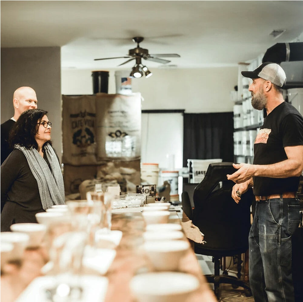 There is a long light wooden table stretching out to the back of the room with several small white bowls lining the sides. On the left of the table two people stand to the side looking the right. On the right side, there is another person talking to the pair on the other side of the table.