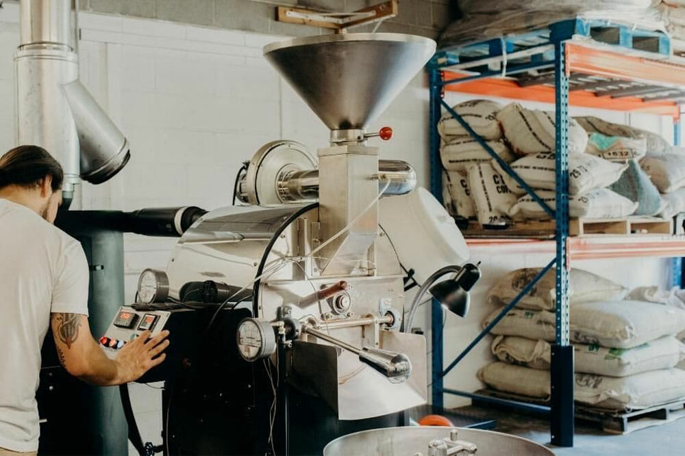 A white roaster is in the center with someone to the right working on the control panel. To the right of the image there are industrial shelves holding green coffee sacks.