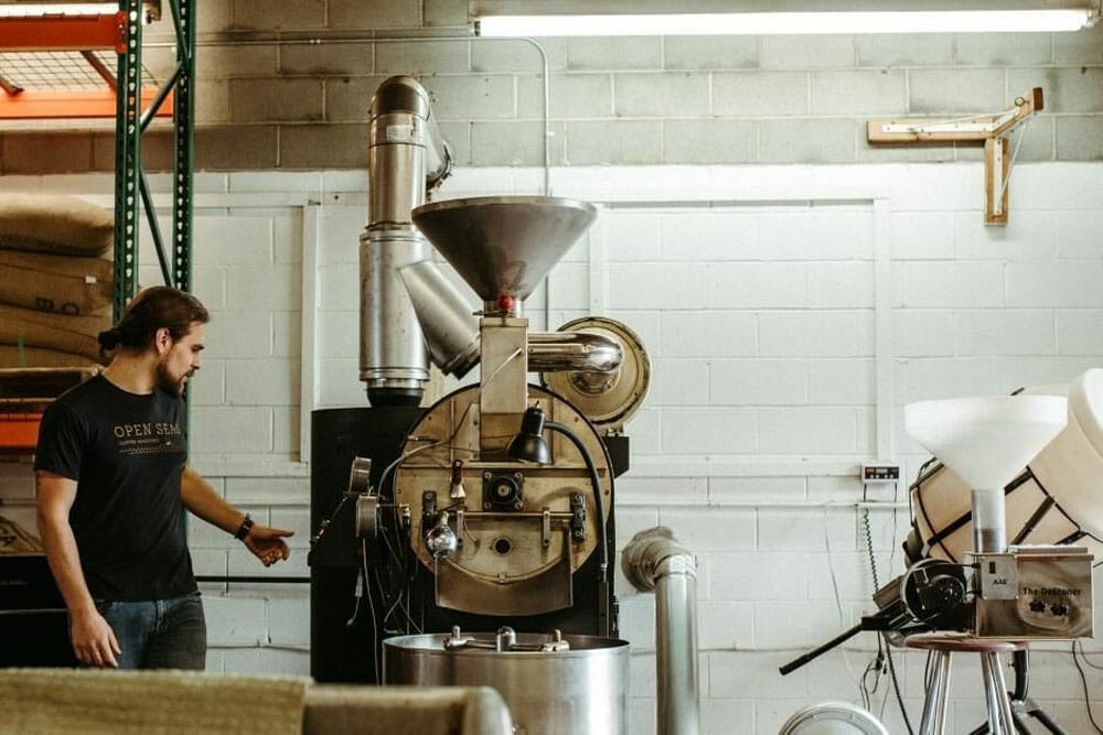Someone standing on the side of a large black roaster. There is a shelf behind them with burlap sacks of green coffee.