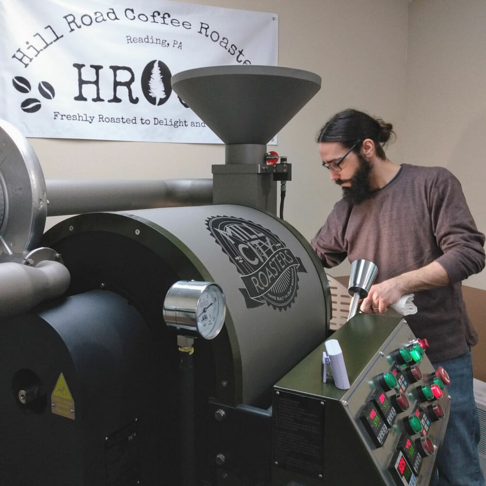 A black, green, and gray colored coffee roaster is positioned diagonally in the frame starting at the bottom left. In front of the roaster, facing the viewer, there is someone opening the cooling tray of the roaster checking on the temperature of the beans. Behind him there is a white sign positioned above that says "Hill Road Coffee Roasters."