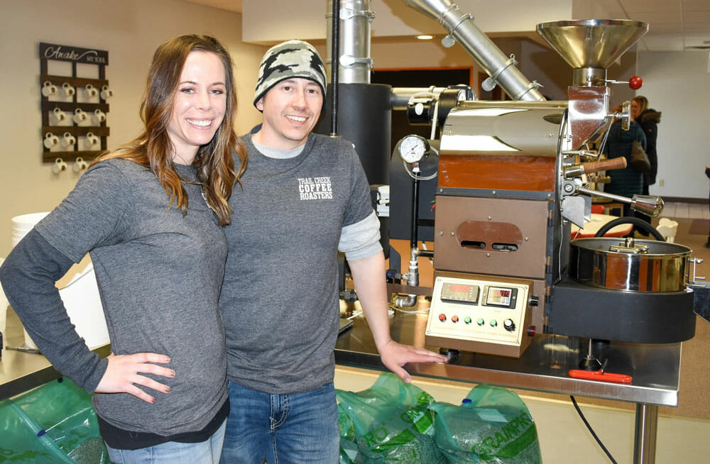 A couple standing together in front of a small roaster. Below them there are bags of green coffee beans.