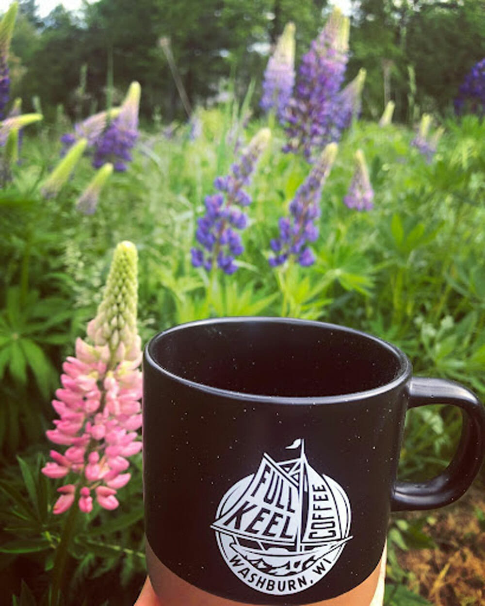 A black mug with a white logo being held up in front of a garden.