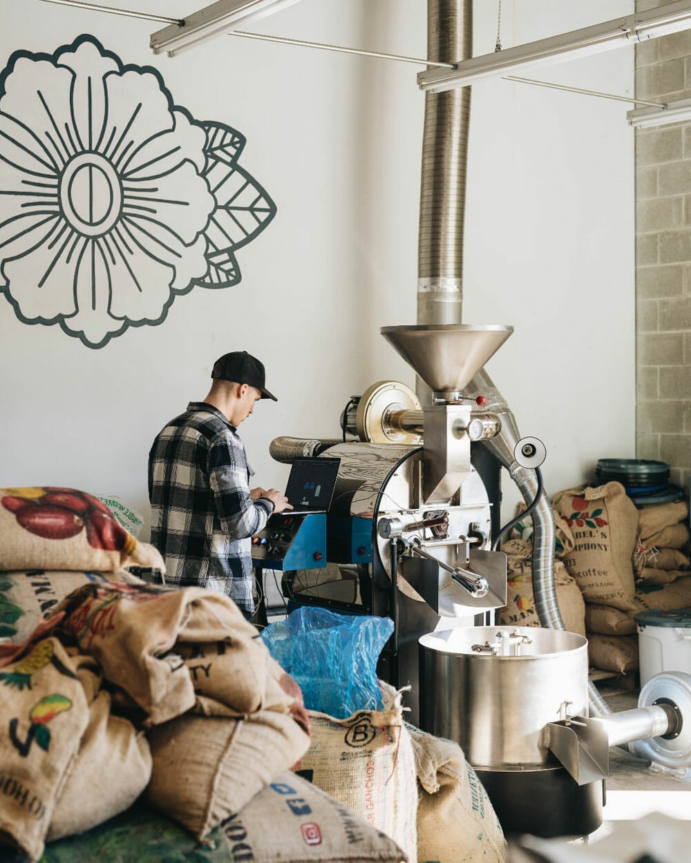Someone standing next to a coffee roaster with burlap green coffee bags surrounding them.
